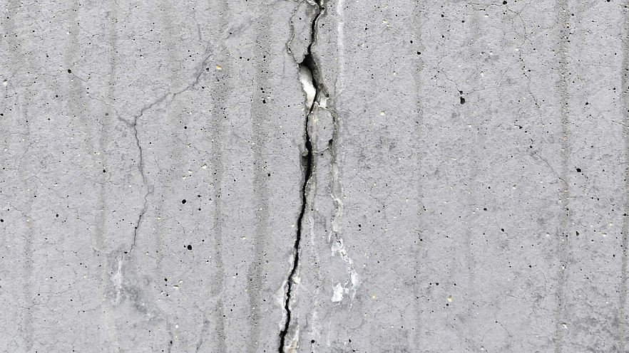 Eine Nahaufnahme aus zeigt eine Betonwand mit einem markanten Riss, der vertikal über die Oberfläche verläuft. Der tiefe und gezackte Riss bildet einen starken Kontrast zum fleckigen Grau des Betons.